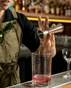 A bartender in a khaki apron pours a clear liquid from a stainless steel jigger into a clear mixing glass containing a pink drink, set on a bar counter with blurred bottles in the background.