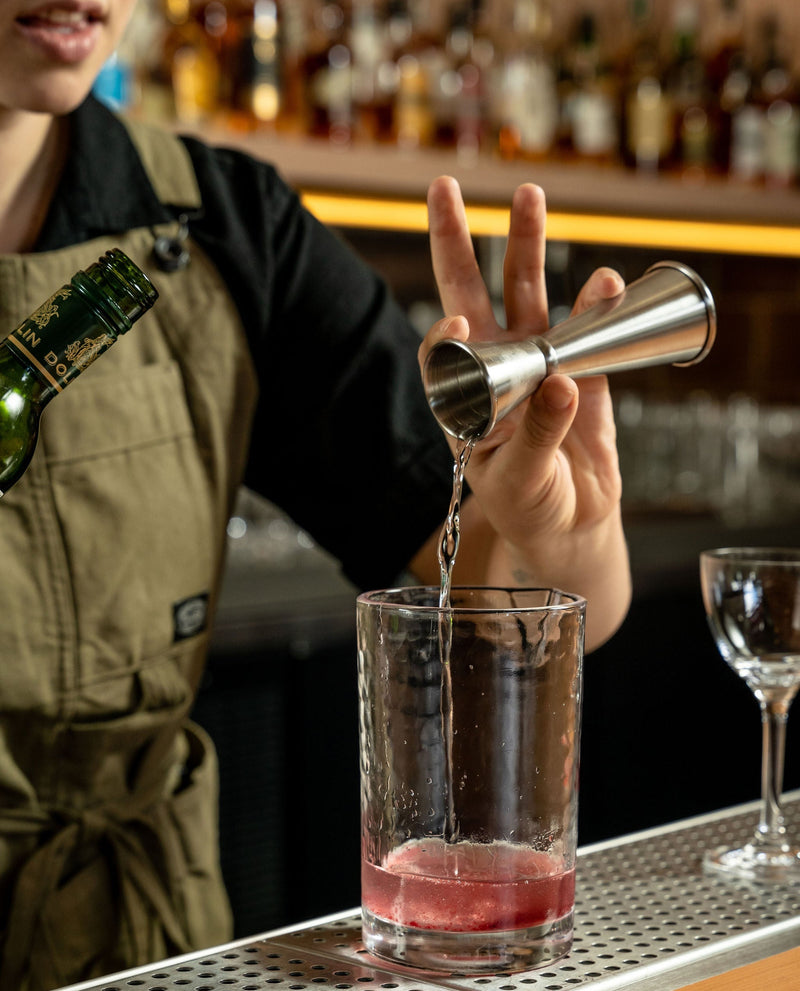 A bartender in a khaki apron pours a clear liquid from a stainless steel jigger into a clear mixing glass containing a pink drink, set on a bar counter with blurred bottles in the background.