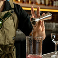 A bartender in a khaki apron pours a clear liquid from a stainless steel jigger into a clear mixing glass containing a pink drink, set on a bar counter with blurred bottles in the background.